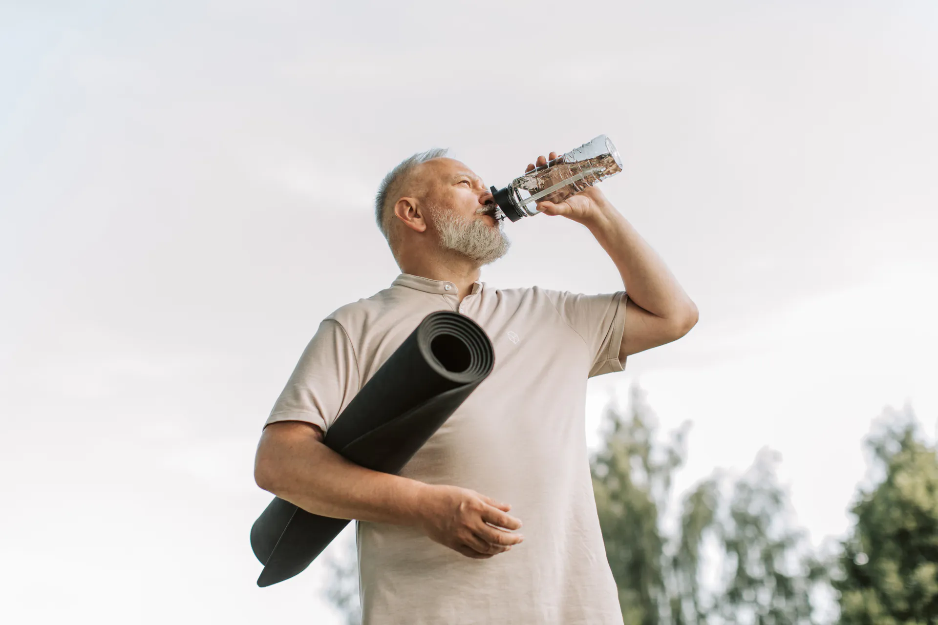Senior man drinking water outdoors with a yoga mat